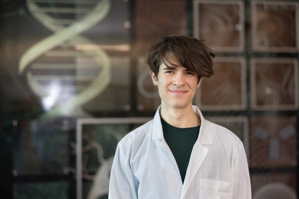 Juliusz Wieckowski smiling and wearing a lab coat in front of the mural in the Science Building.